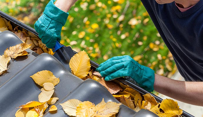A person wearing teal work gloves using a small scoop to remove fallen autumn leaves and debris from a black residential gutter system