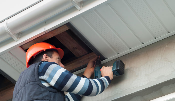 A close-up view of a technician using a power drill to secure white soffit panels to the underside of a roof overhang