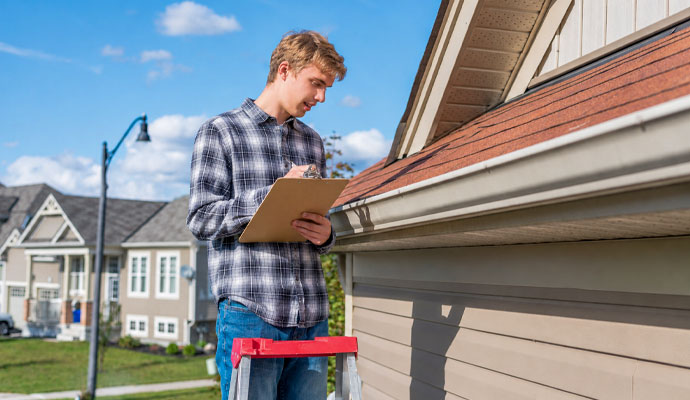 Technician on a ladder with a clipboard inspecting roof gutters