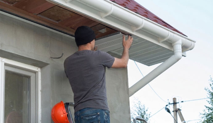 A technician on a ladder replacing white perforated soffit panels under the eaves of a residential home