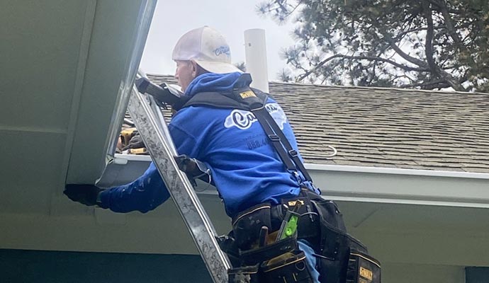 A technician on a ladder installing a white seamless gutter system on a residential home with a shingled roof