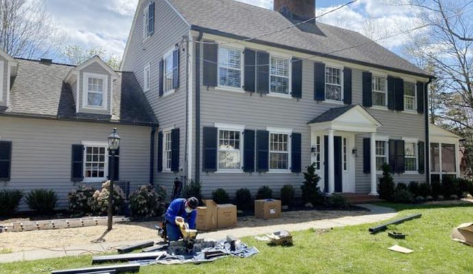 A professional technician in a blue uniform working on gutter components in front of a large two-story grey residential home
